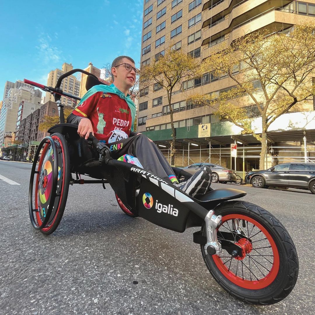 A child sits in an adapted running vehicle, a essentially three-wheel streamlined carriage, on an empty New York City street.  The viewpoint is from close to the ground, looking up at the child.  The child is wearing a red racing jersey, a mint green cape, a NYC Marathon race number, and a determined expression as they look forward.  Behind them stand a number of high-rise buildings and some parked cars along the side of the street, and a mostly-clear blue sky is visible between buildings.
